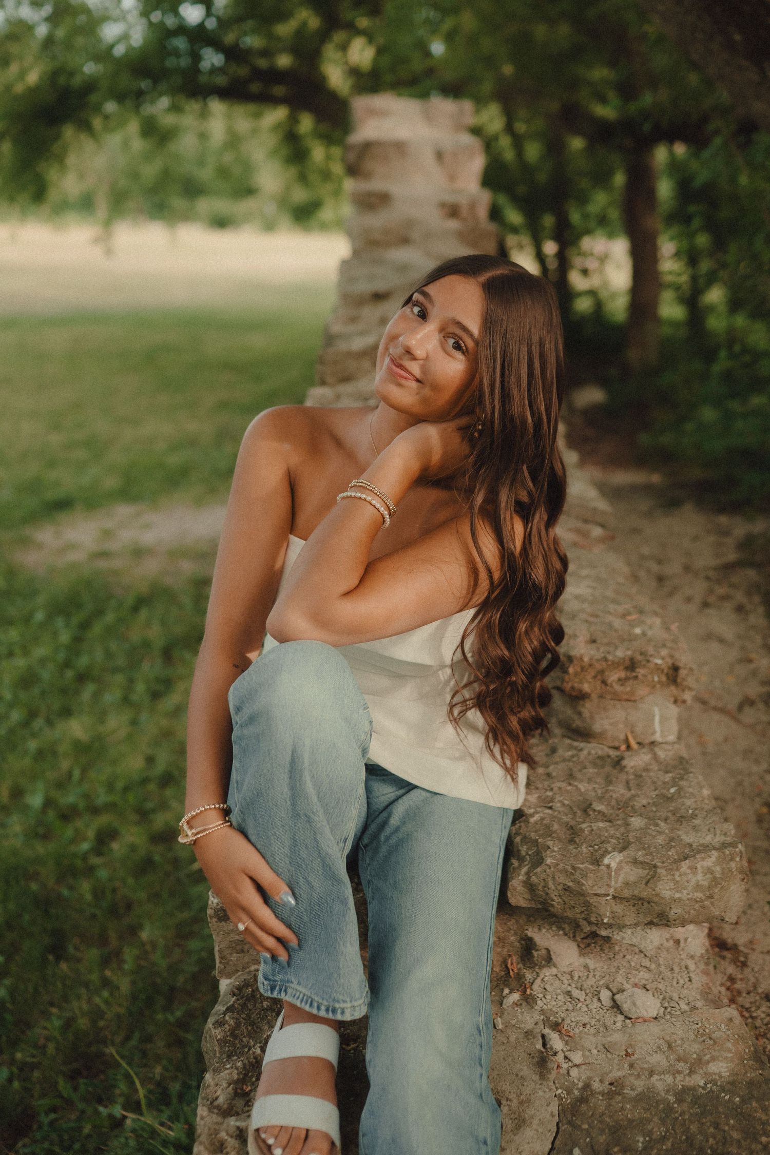 Young woman with long wavy hair sits on stone wall in white top and jeans, smiling in a lush green park.