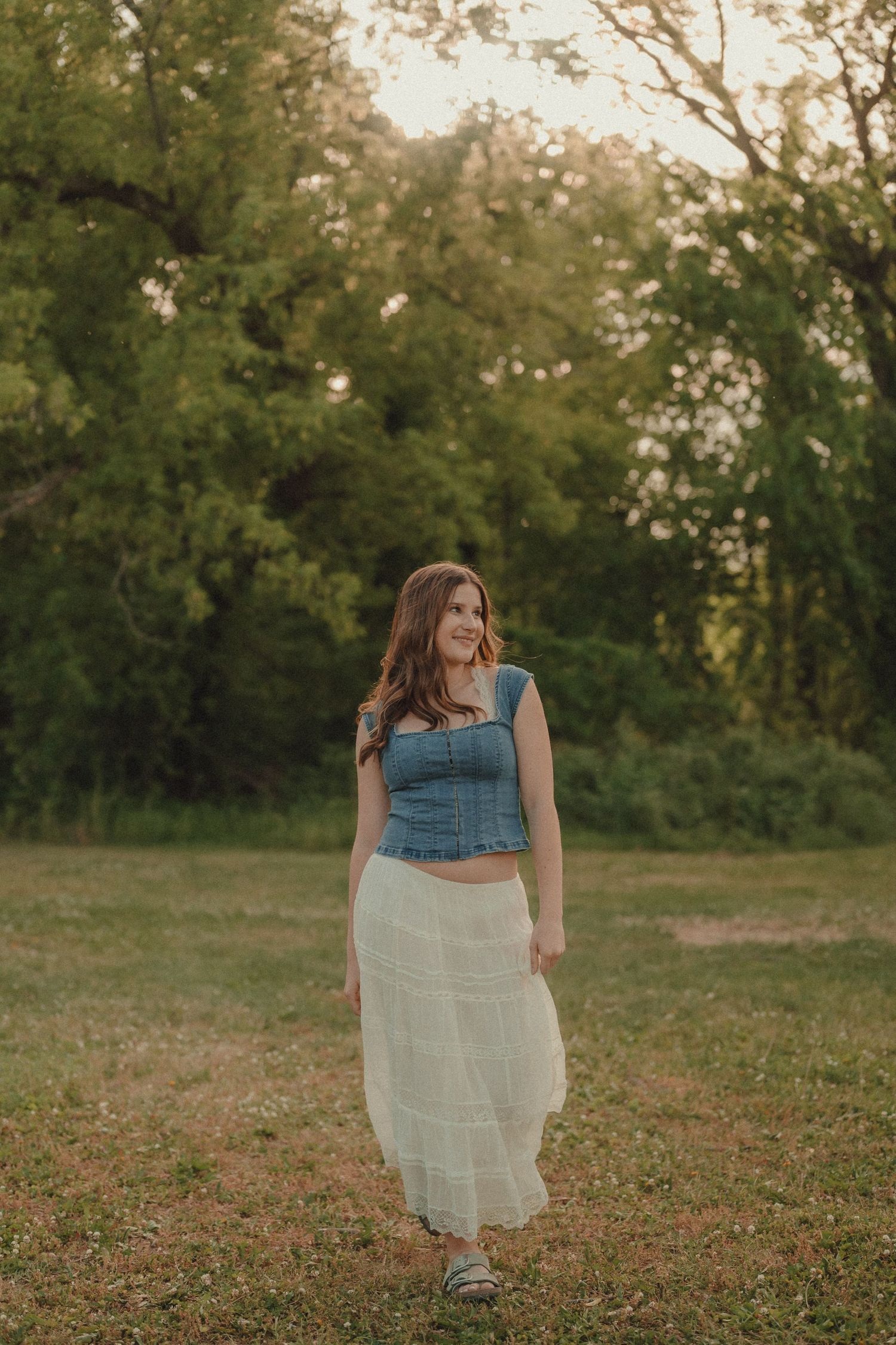 Young woman in denim crop top and white maxi skirt standing in a sunlit green field at golden hour.