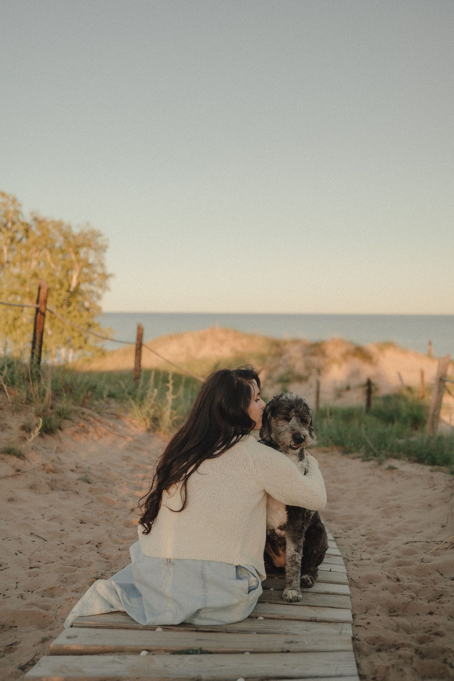 Woman in white sweater hugs curly dog on wooden boardwalk overlooking sandy beach at sunset.