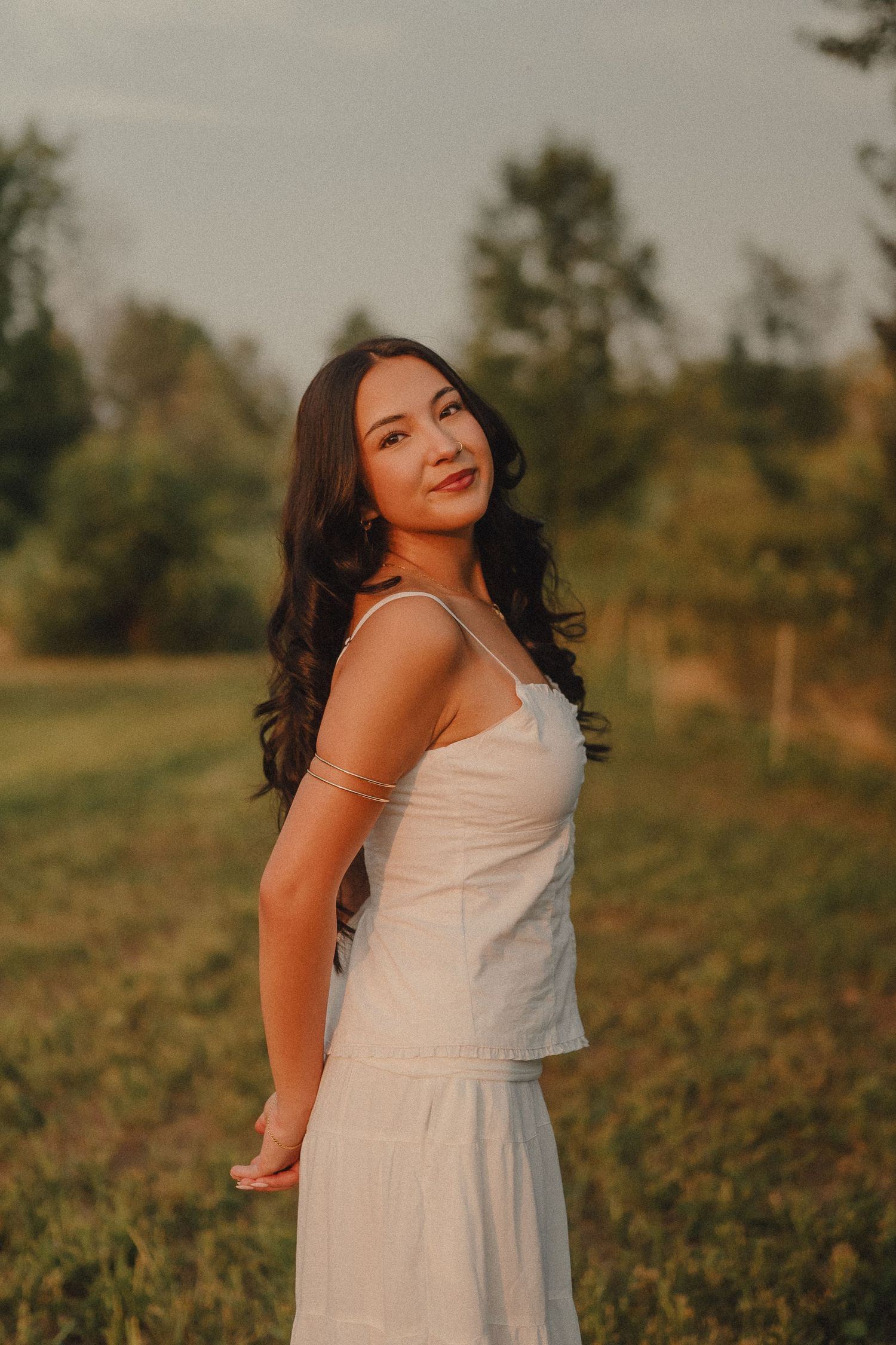 Beautiful brunette woman in white sundress smiling outdoors in a green field at sunset.