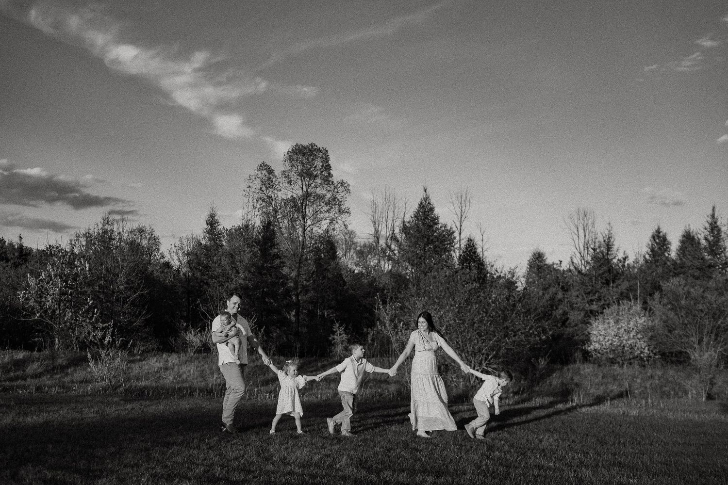 Black and white photo of a family holding hands in a field with evergreen trees at dusk.