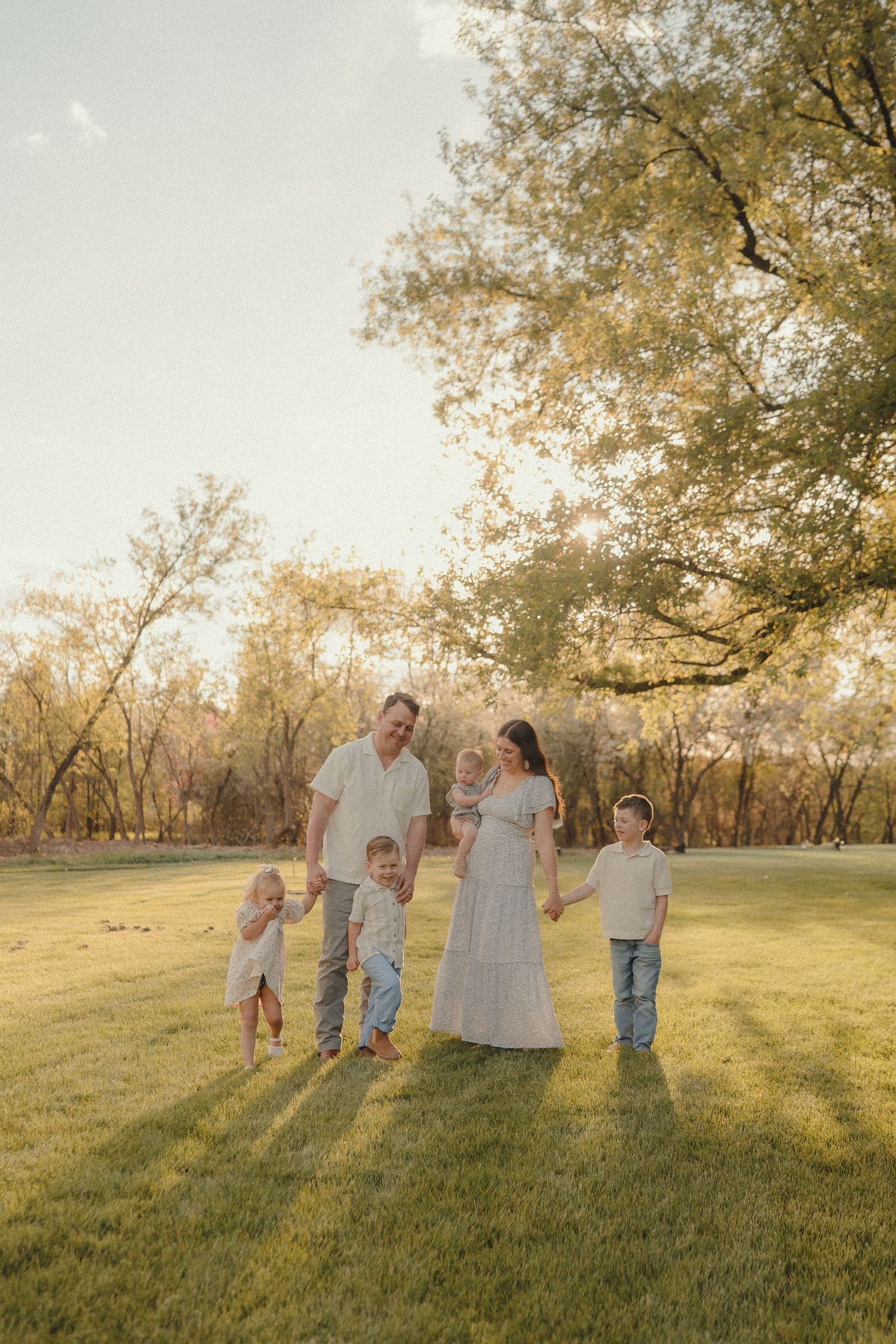 Family of six walking hand-in-hand across a sunlit green meadow surrounded by autumn trees.