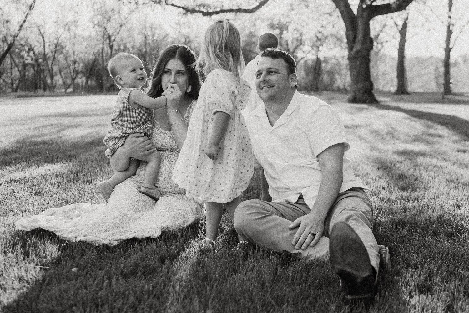 Black and white family photo of parents with two young children sitting on grass in a park.