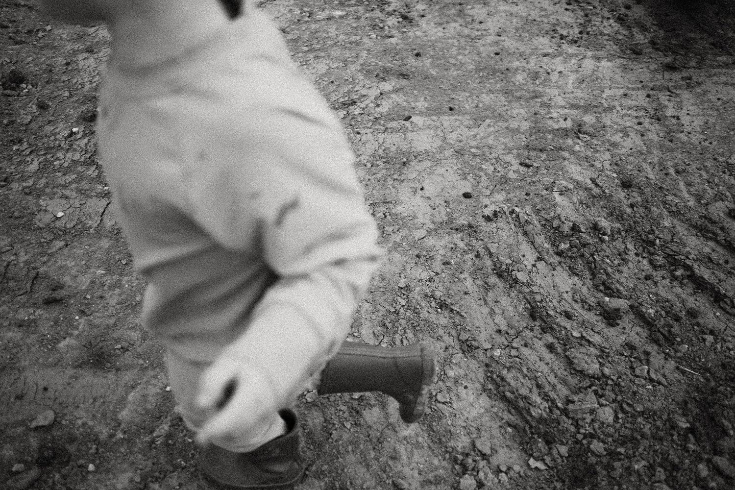 Black and white photo of a person in work clothes holding a tool, walking on muddy ground outdoors.