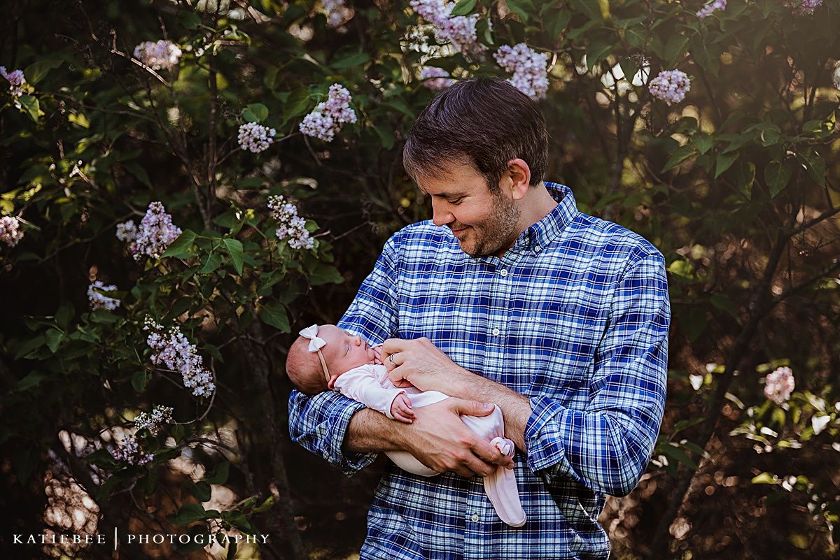 A newborn baby wrapped in white rests in caring arms against a backdrop of flowering bushes.