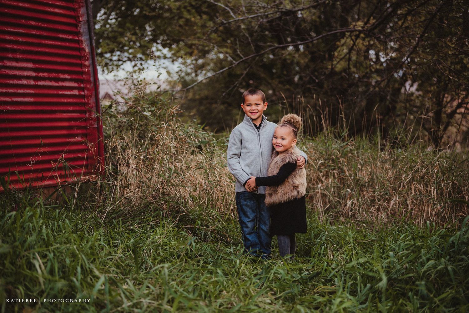 Two children playing together in tall grass near a red barn structure.