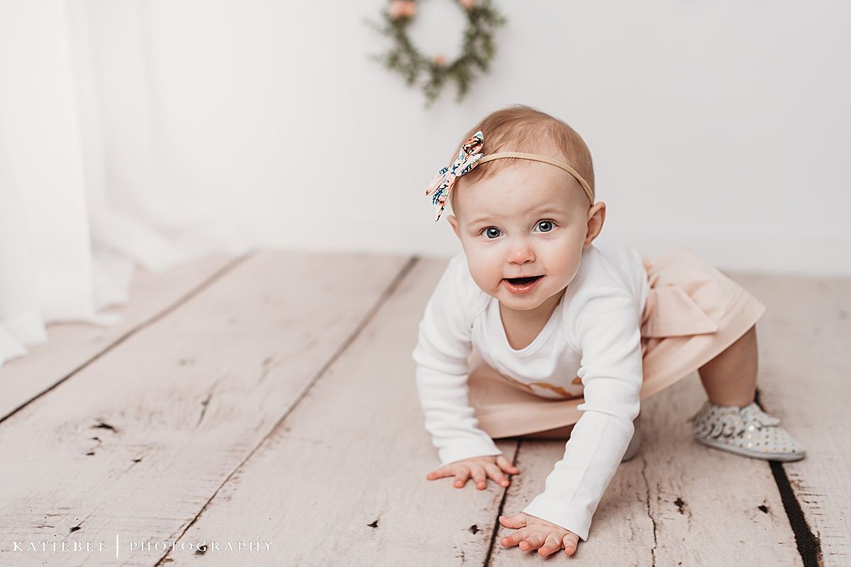 Baby wearing white top and pink skirt sitting by cake and floral wreaths.