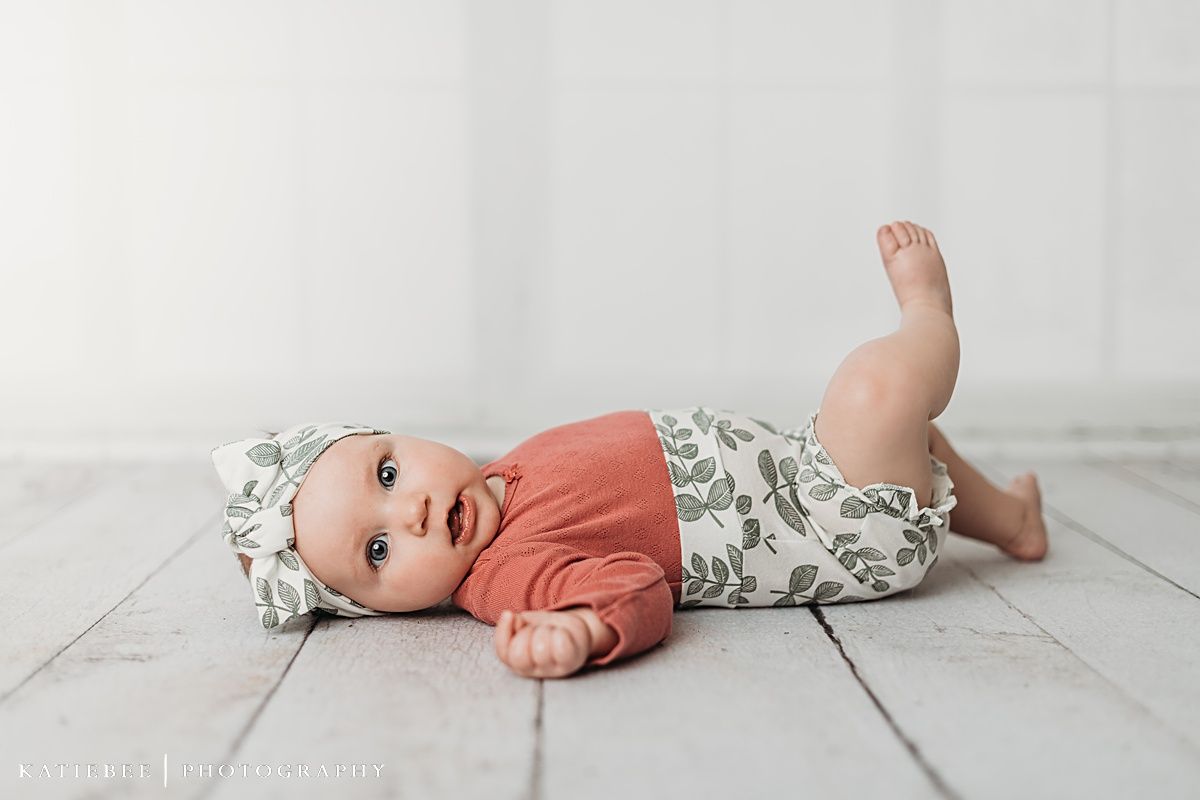 Happy baby in coral top and floral outfit lying on white wooden floor.