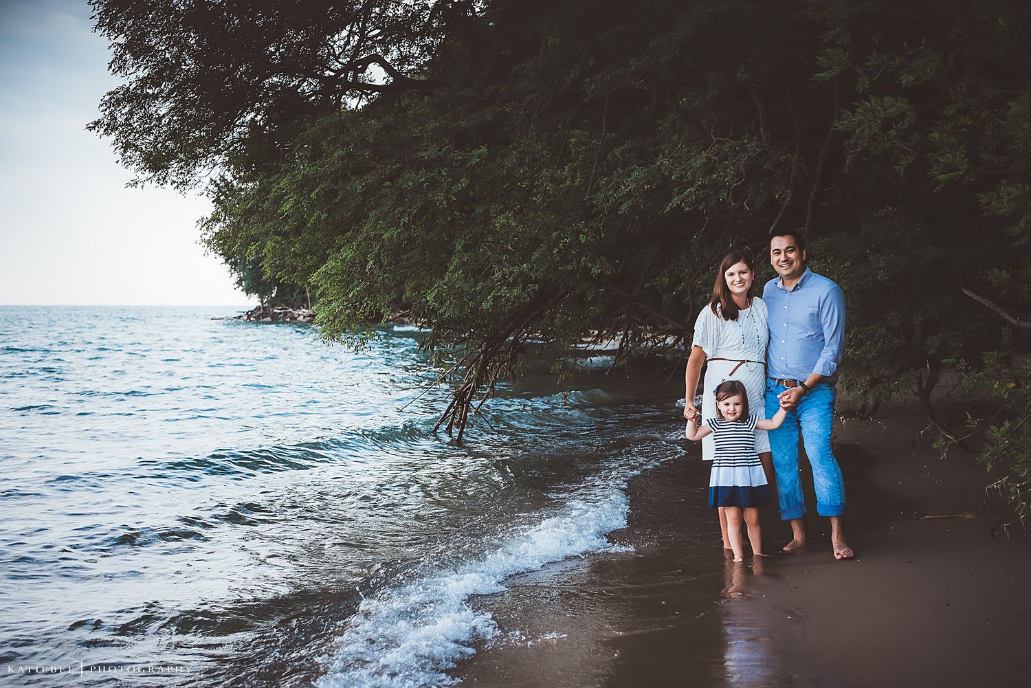 A couple walks along a scenic beach shoreline with trees and waves in the background.