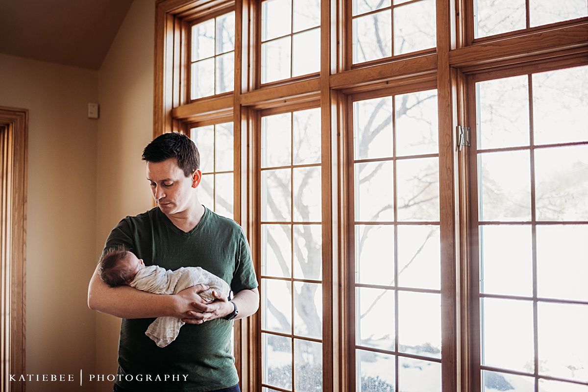 Parent in green shirt holds sleeping newborn by sunlit windows.