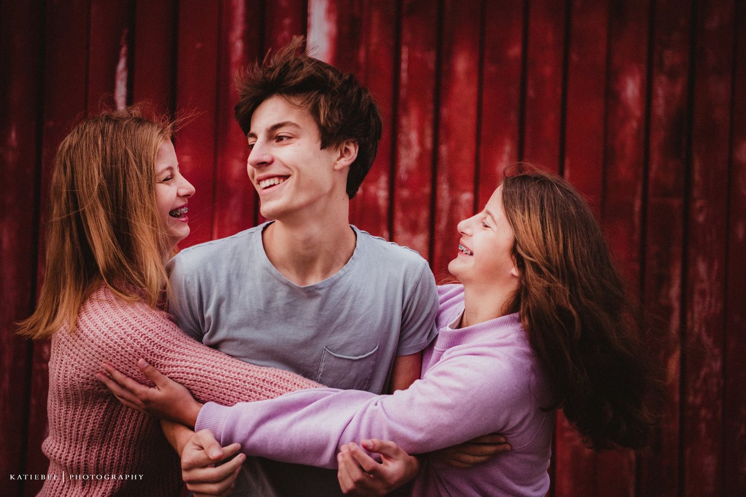 Three friends laughing together in front of a red barn wall during a casual photo session.