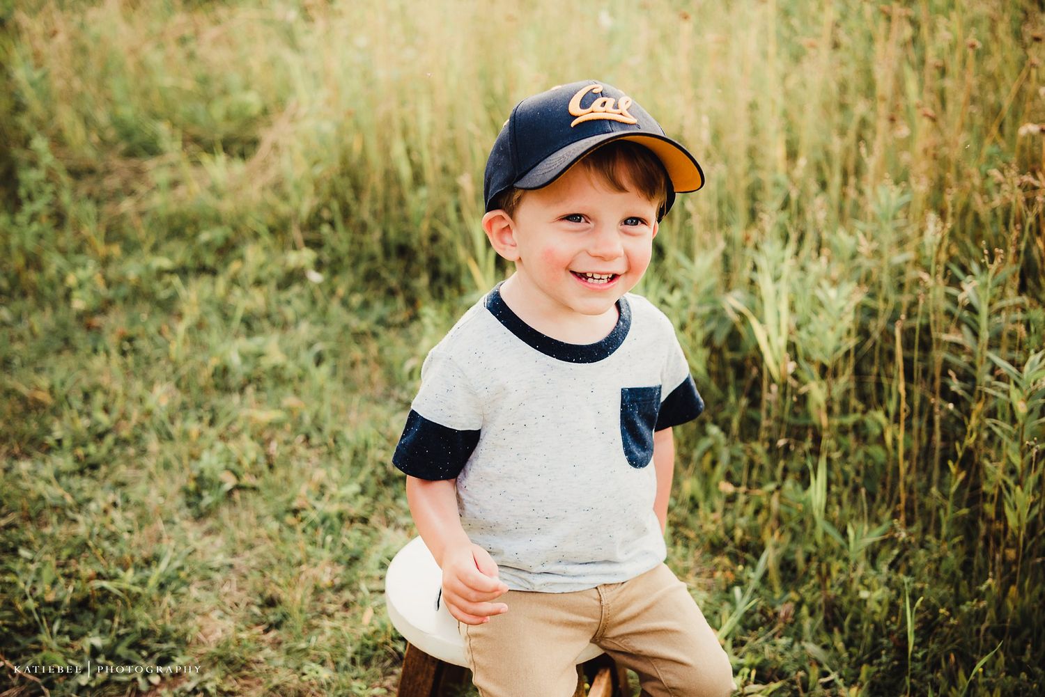 Young boy wearing a baseball cap smiles brightly while playing in tall grass.