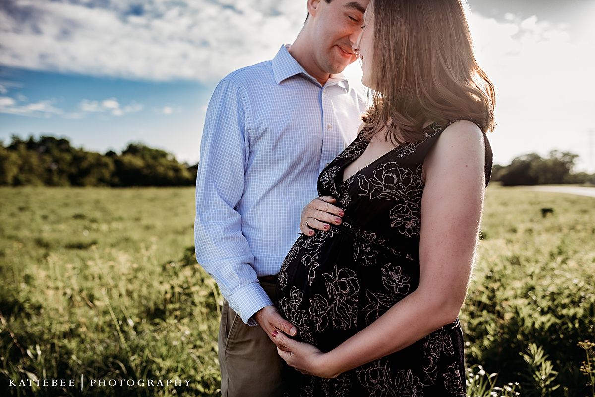 An intimate maternity portrait of a couple in a sunny field with dramatic clouds.