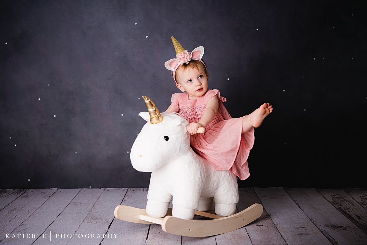 A toddler in a pink dress rides a white rocking unicorn toy against a dark background.