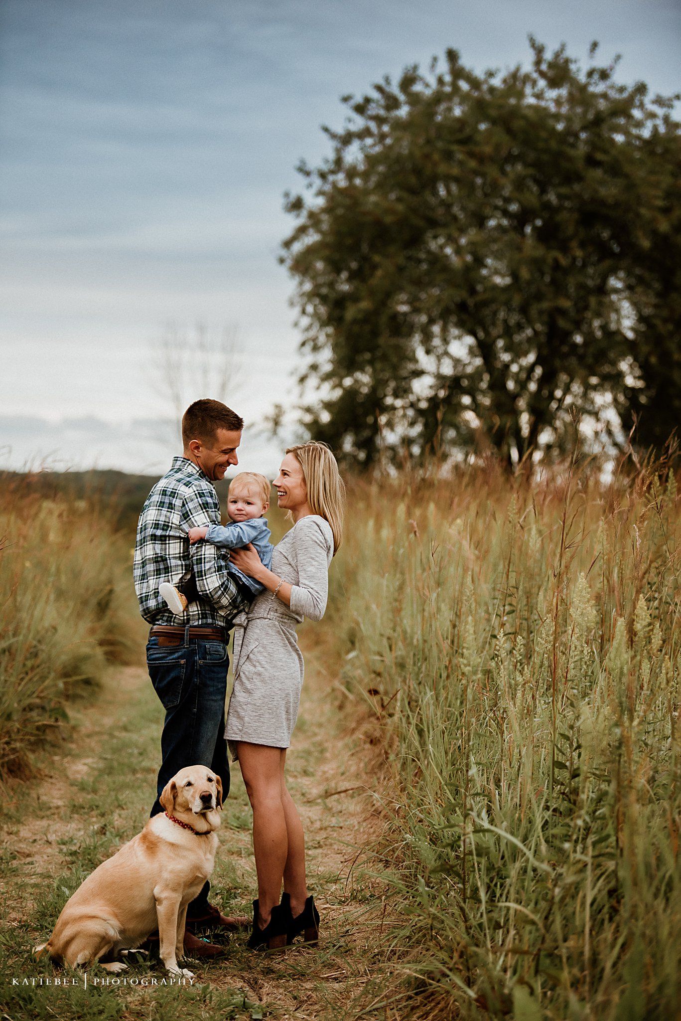 Couple stands on nature path with their labrador as tall grasses and trees surround them.