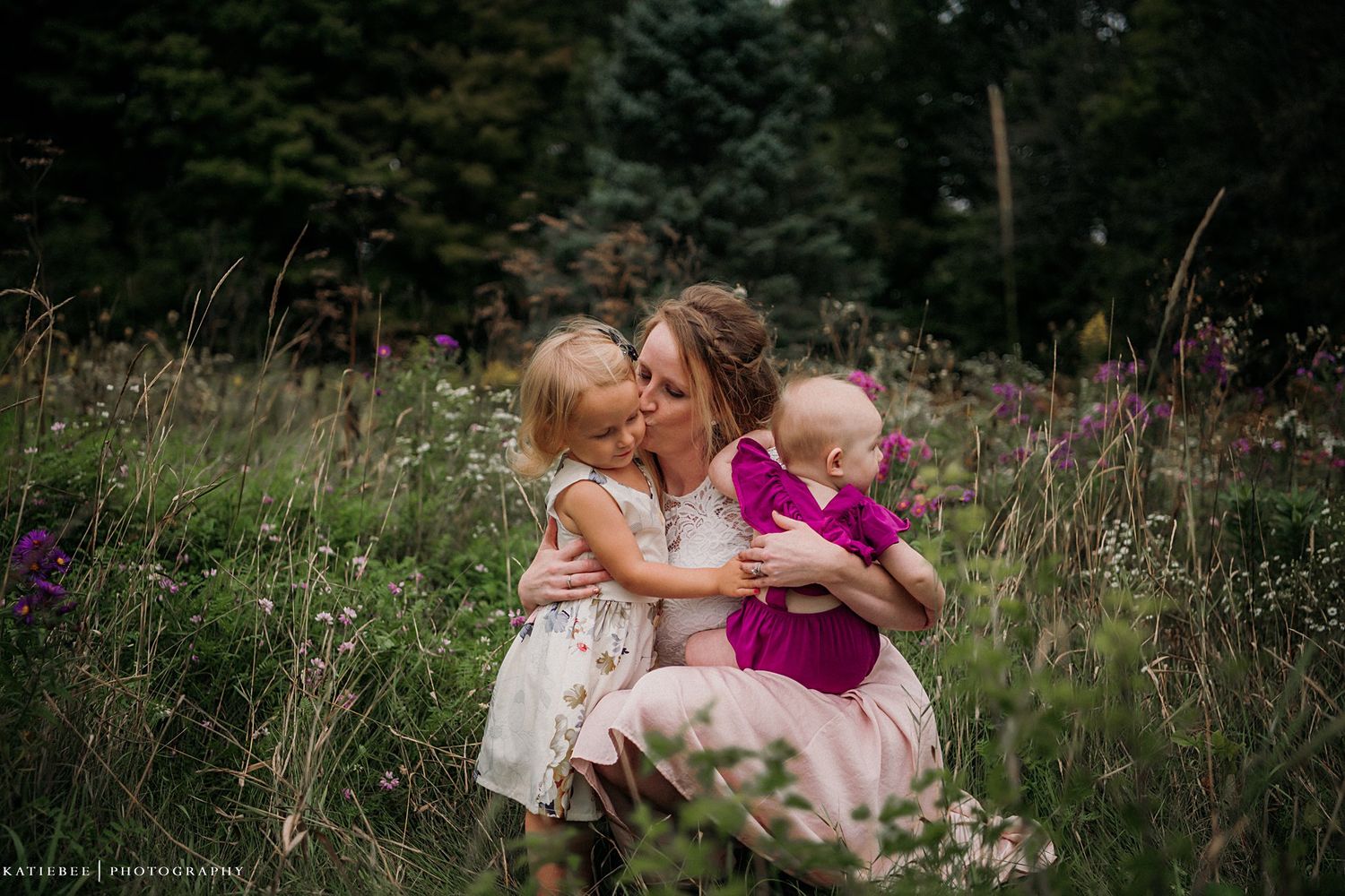 Three figures share an intimate moment together among purple wildflowers in a summer meadow.