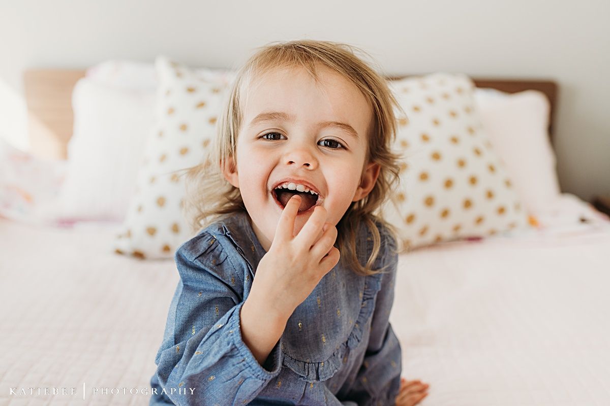 Joyful toddler in denim dress laughs while sitting on bed with polka dot pillows.