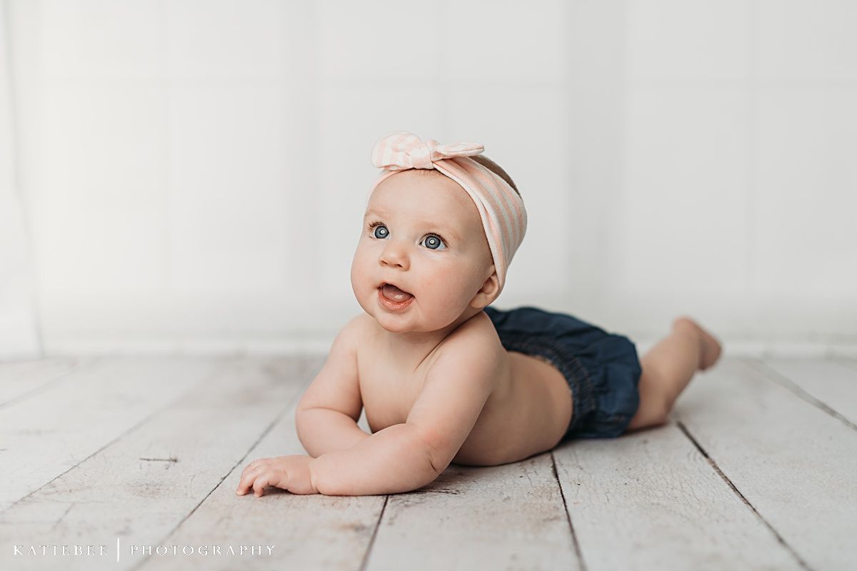 Baby in blue shorts lying on white wooden floor with headband during minimalist photo session.