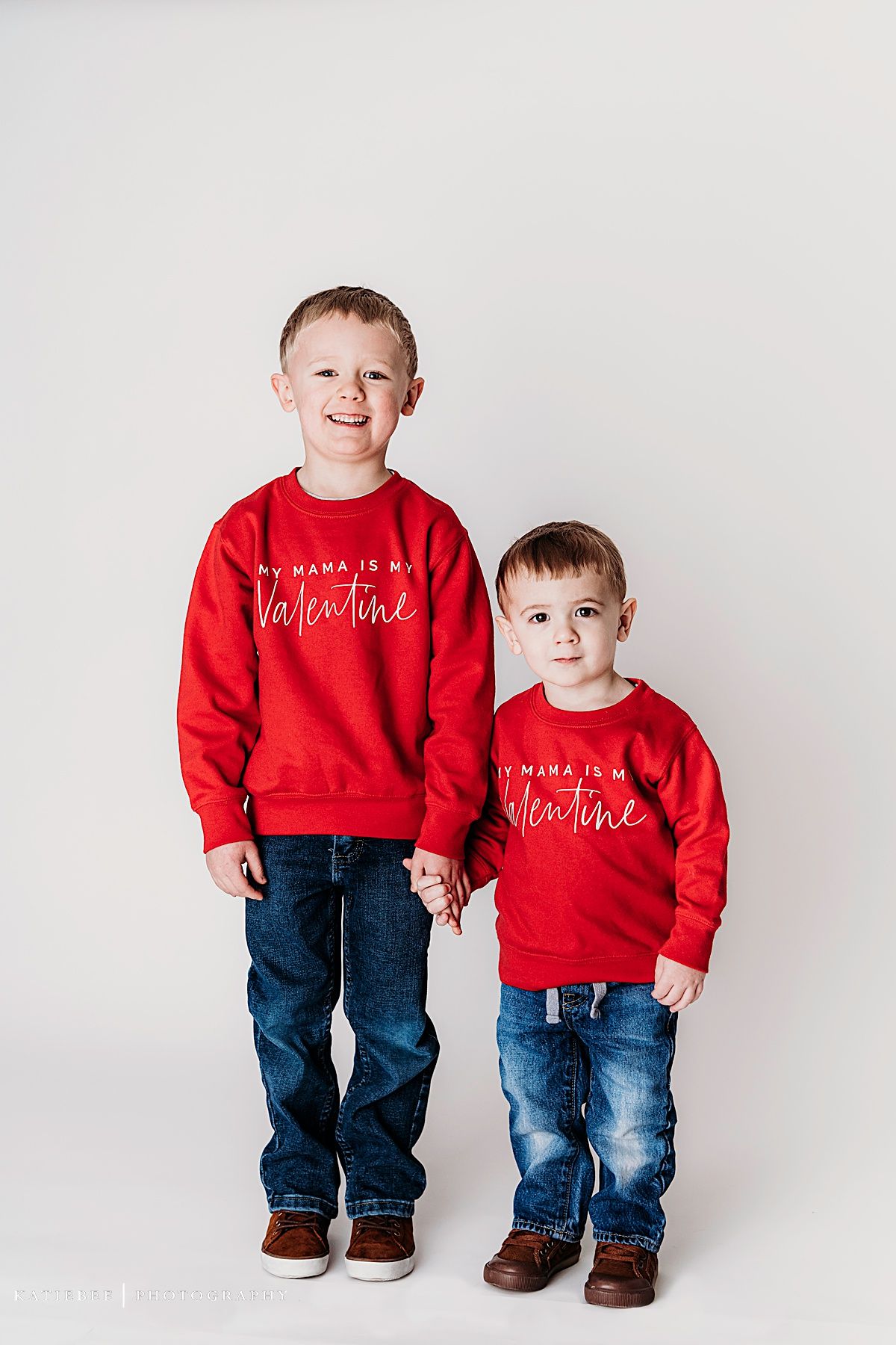 Two children wearing matching red Valentine's sweatshirts and jeans stand against a white backdrop.
