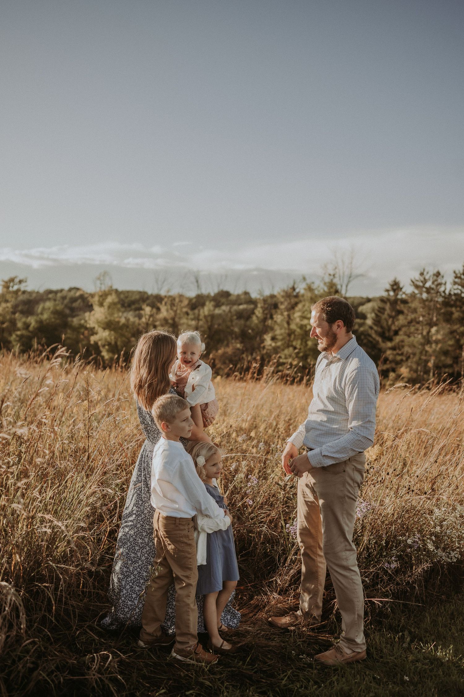 Family gathering in sunlit meadow with mountain landscape and pine trees in background.