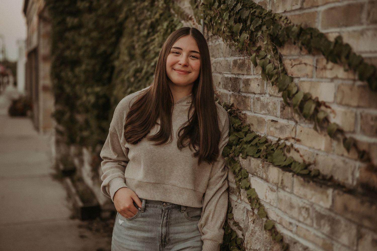 Person in beige sweater standing against ivy-covered brick wall outdoors.