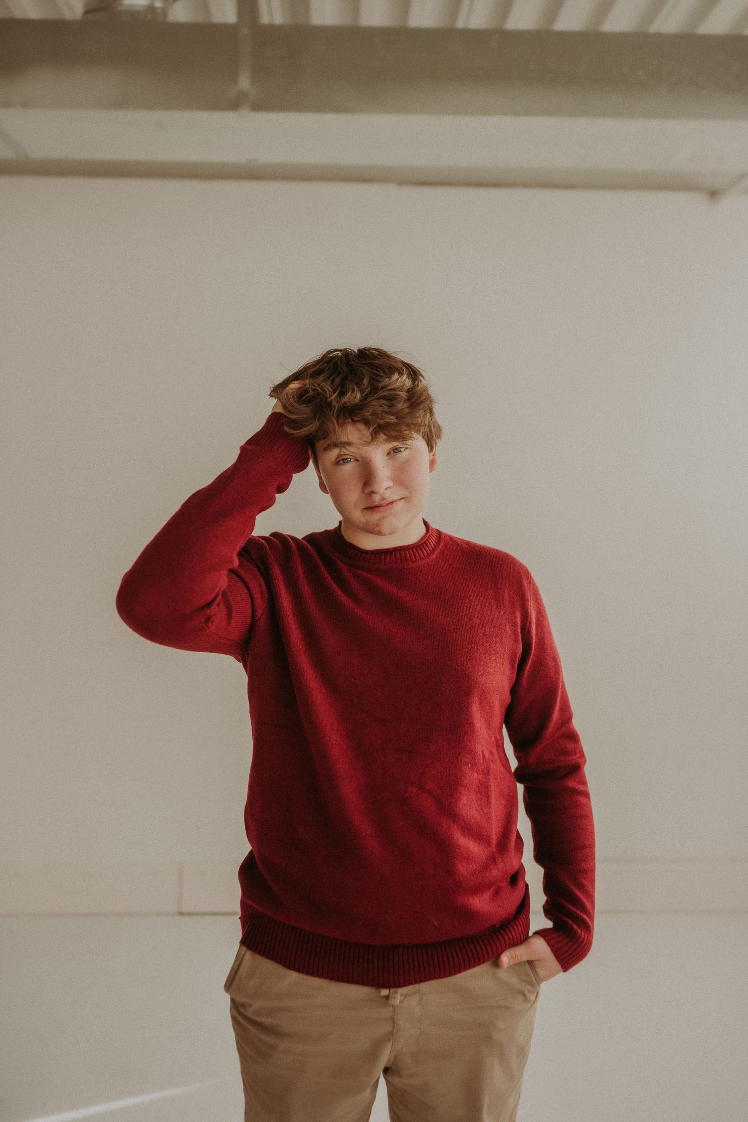 A casual portrait in warm tones featuring a person in a red sweater with hand raised to head.