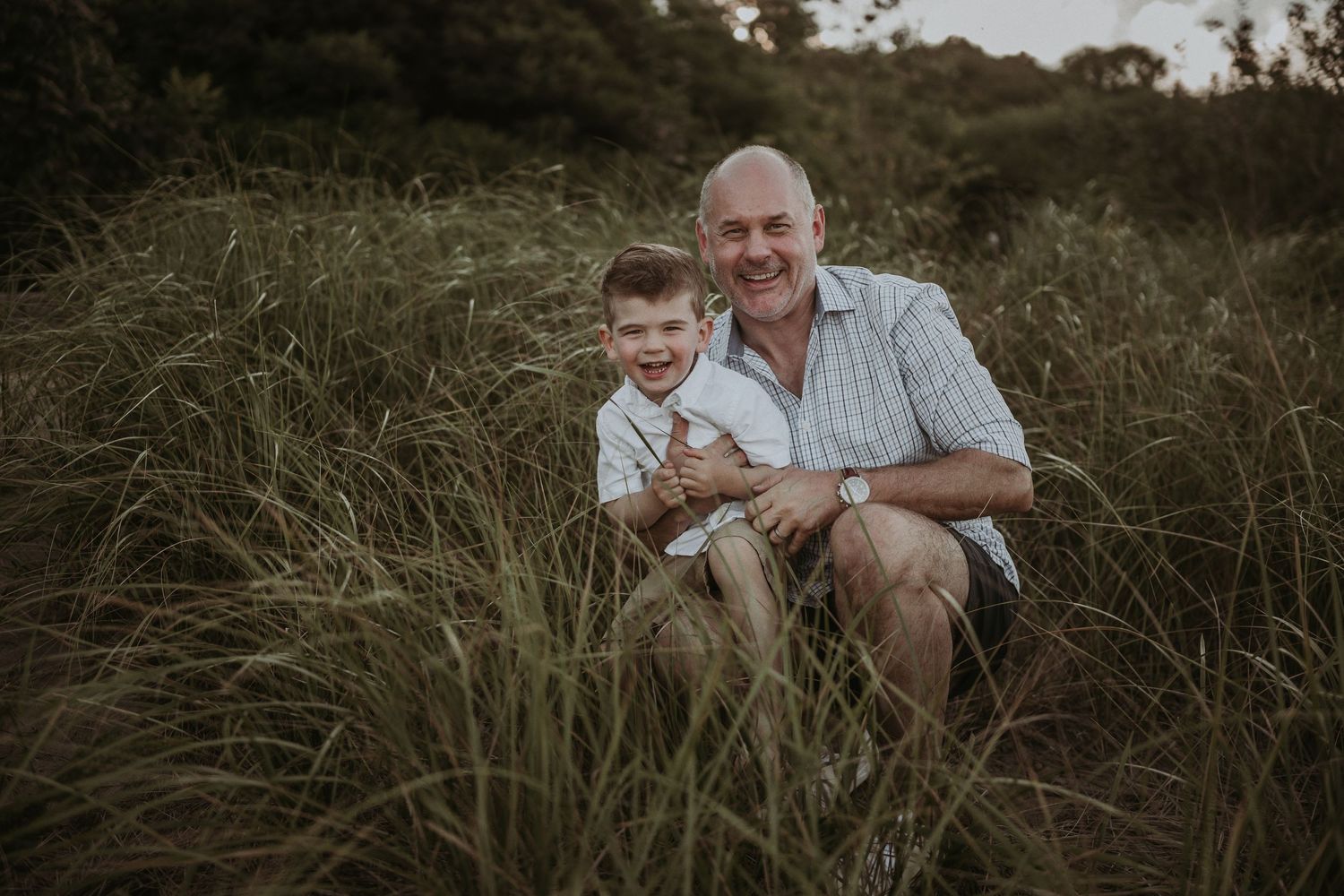 An intimate portrait of two people sitting together in tall grass during sunset.