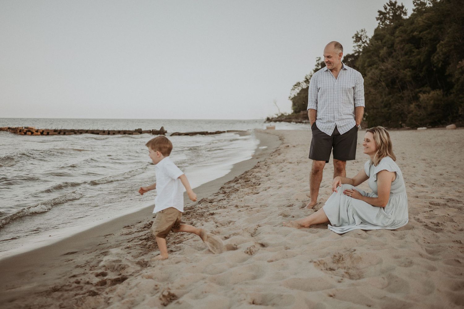 Child plays in the sand while adults relax on a scenic beach with trees.