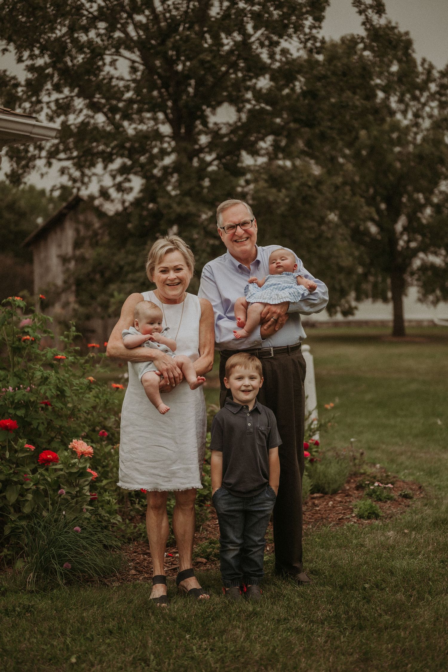 Family portrait in garden setting with red flowers and rustic barn in background.