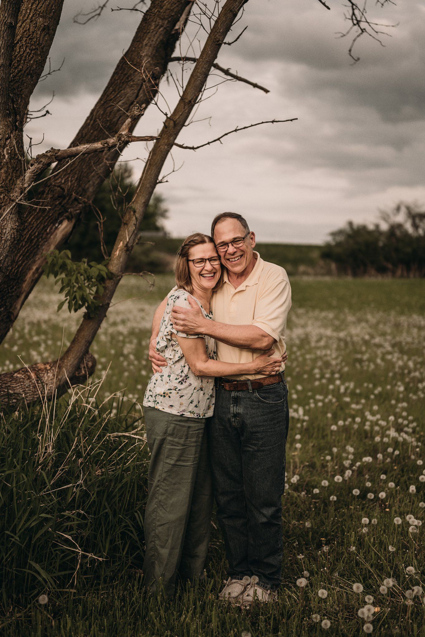 Couple embracing in wildflower field beneath dramatic cloudy sky and leaning tree.