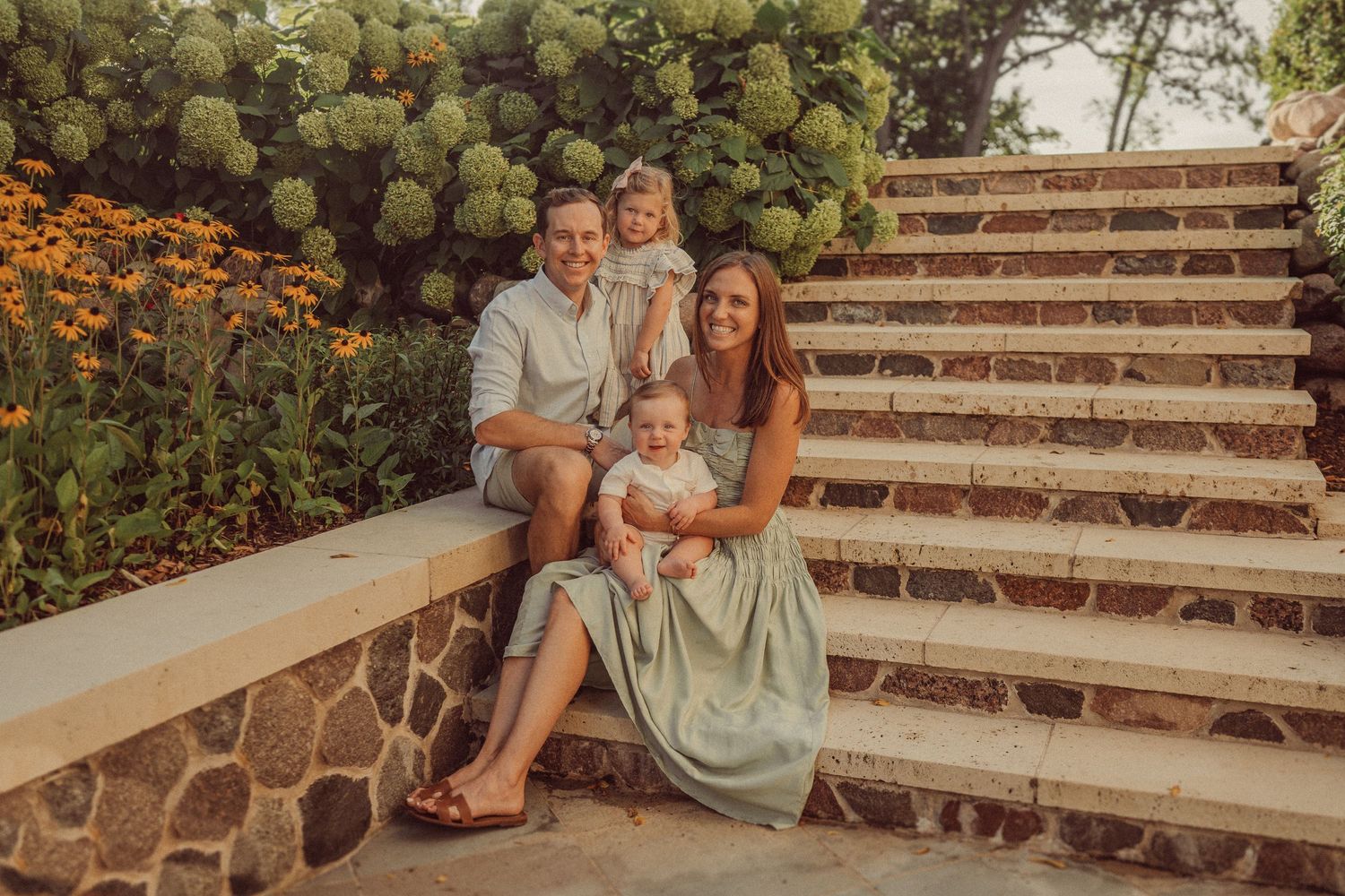 A family sits together on stone stairs surrounded by vibrant hydrangeas and orange flowers in a garden setting.