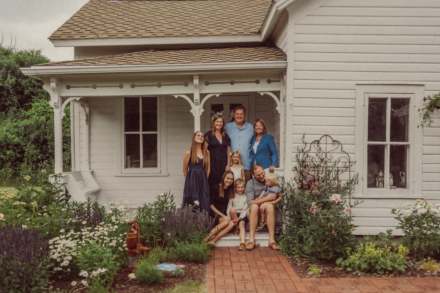 A group poses together on the front porch steps of a classic white farmhouse surrounded by blooming flowers.
