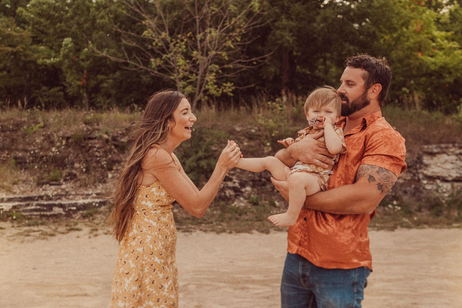 A family shares a playful moment on a sandy riverbank during golden hour.