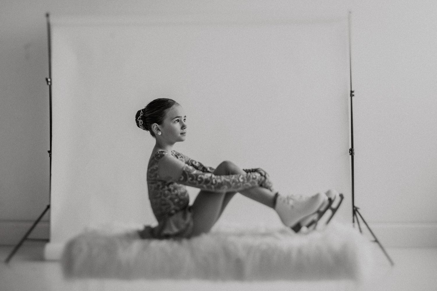 Figure skater sits gracefully on white fur surface in dramatic black and white studio shot.