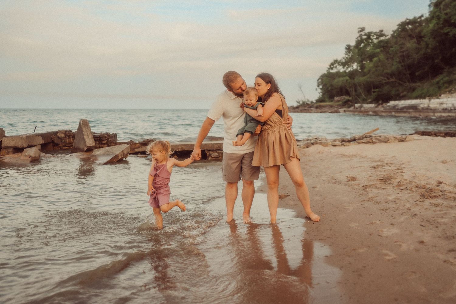 A family plays and embraces at the water's edge during golden hour on a rocky beach.