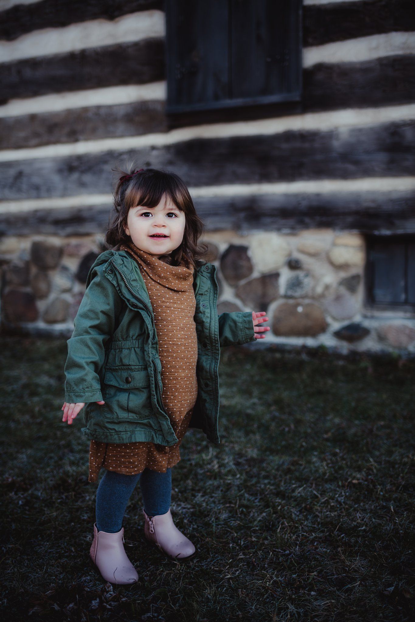 A toddler in a green jacket and brown dress stands near a rustic stone wall.