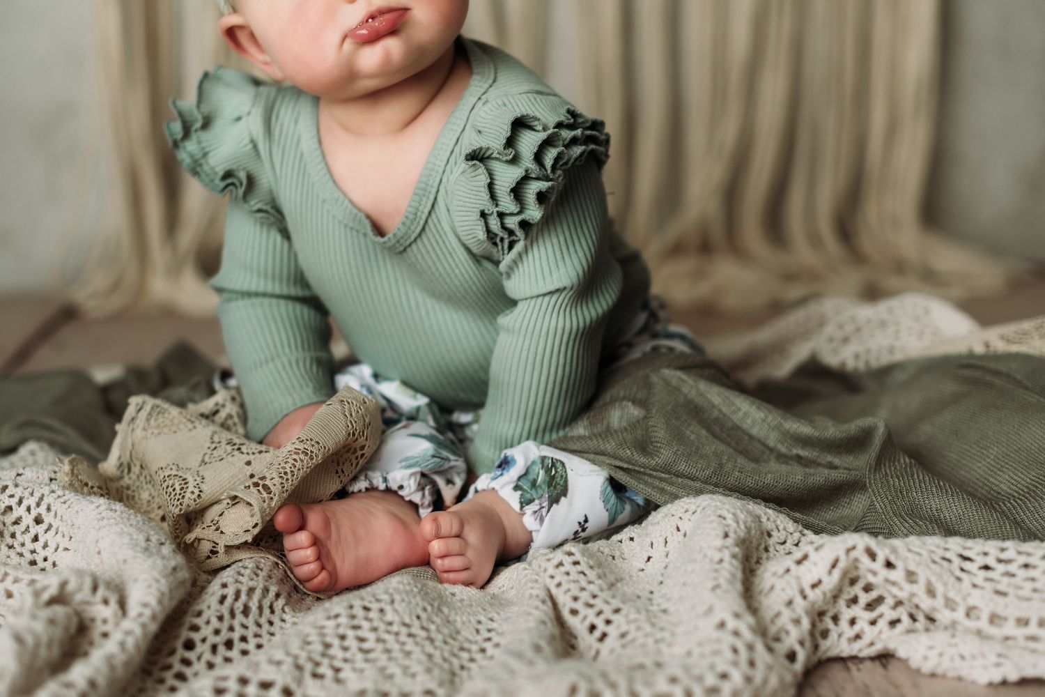 Close up of baby wearing ruffled sage green top sitting on crocheted blanket.