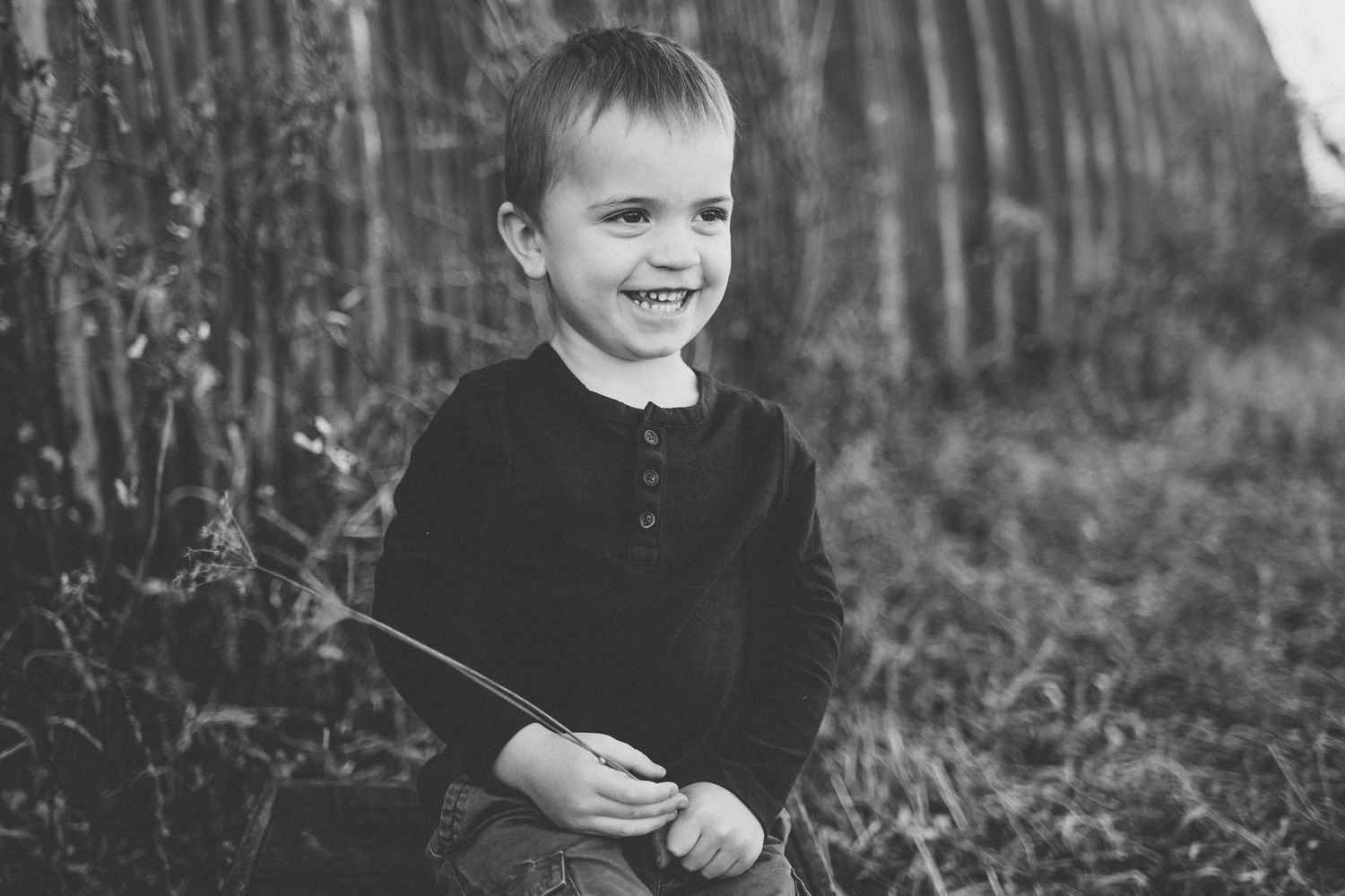 Black and white portrait of a young child holding a stick while standing outdoors near a rustic wall.