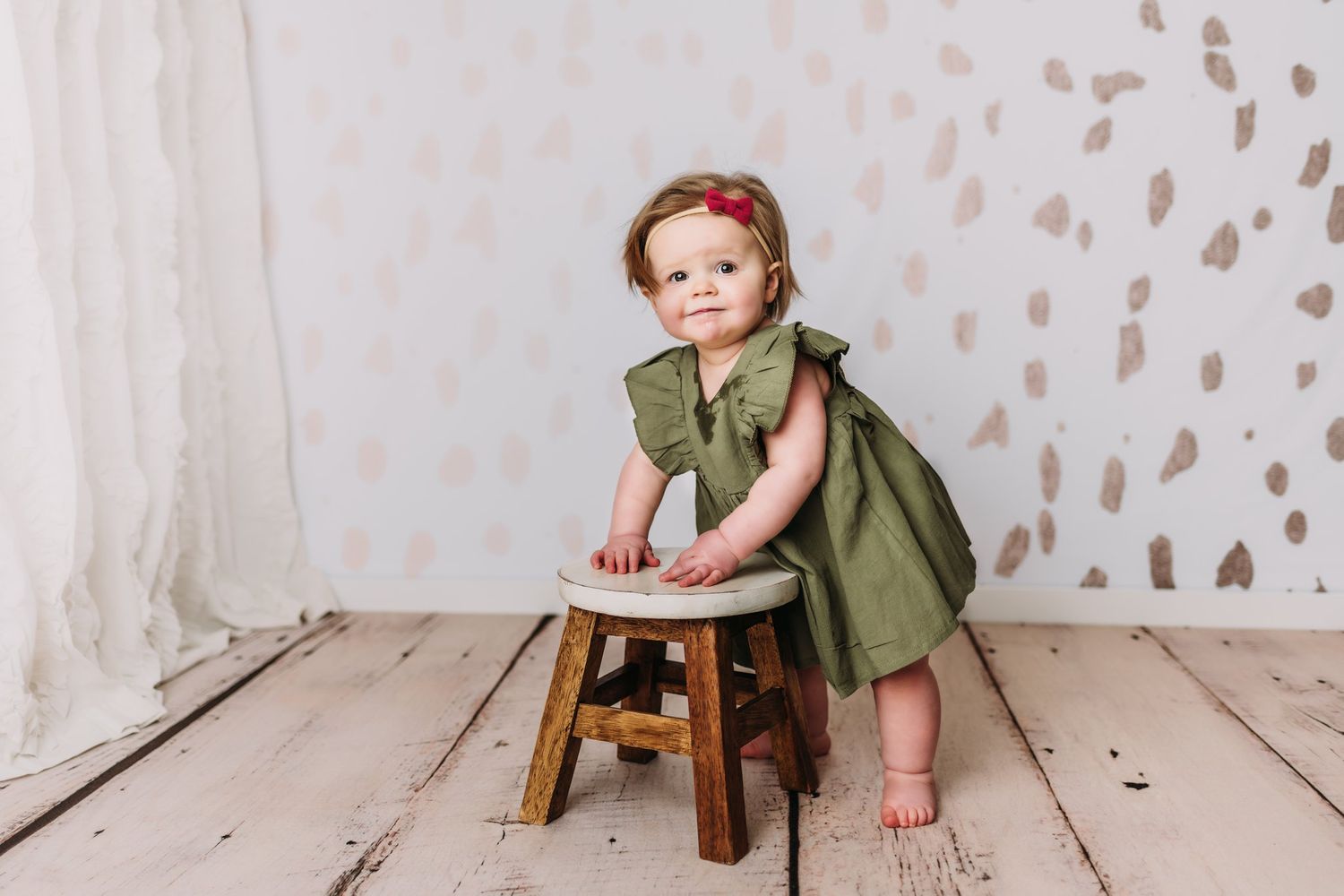 Sweet portrait with wooden stool and olive dress against textured white backdrop.