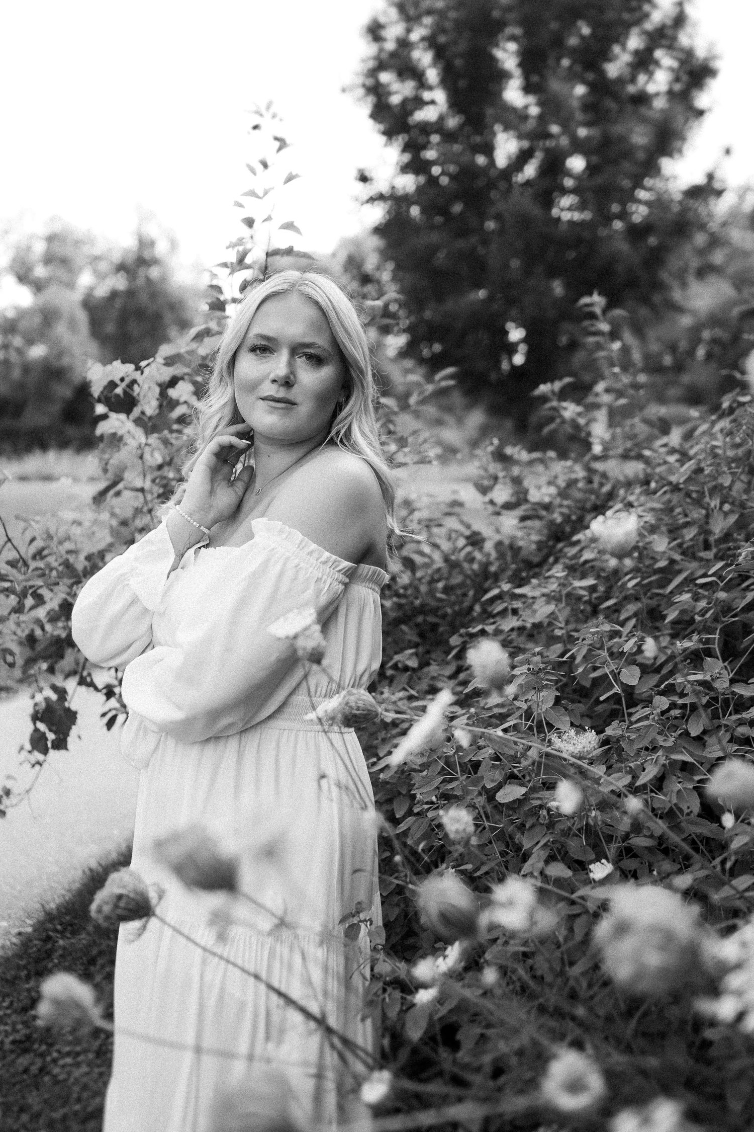 Artistic black and white portrait series showing a woman in a white dress posing elegantly among flowering bushes outdoors.