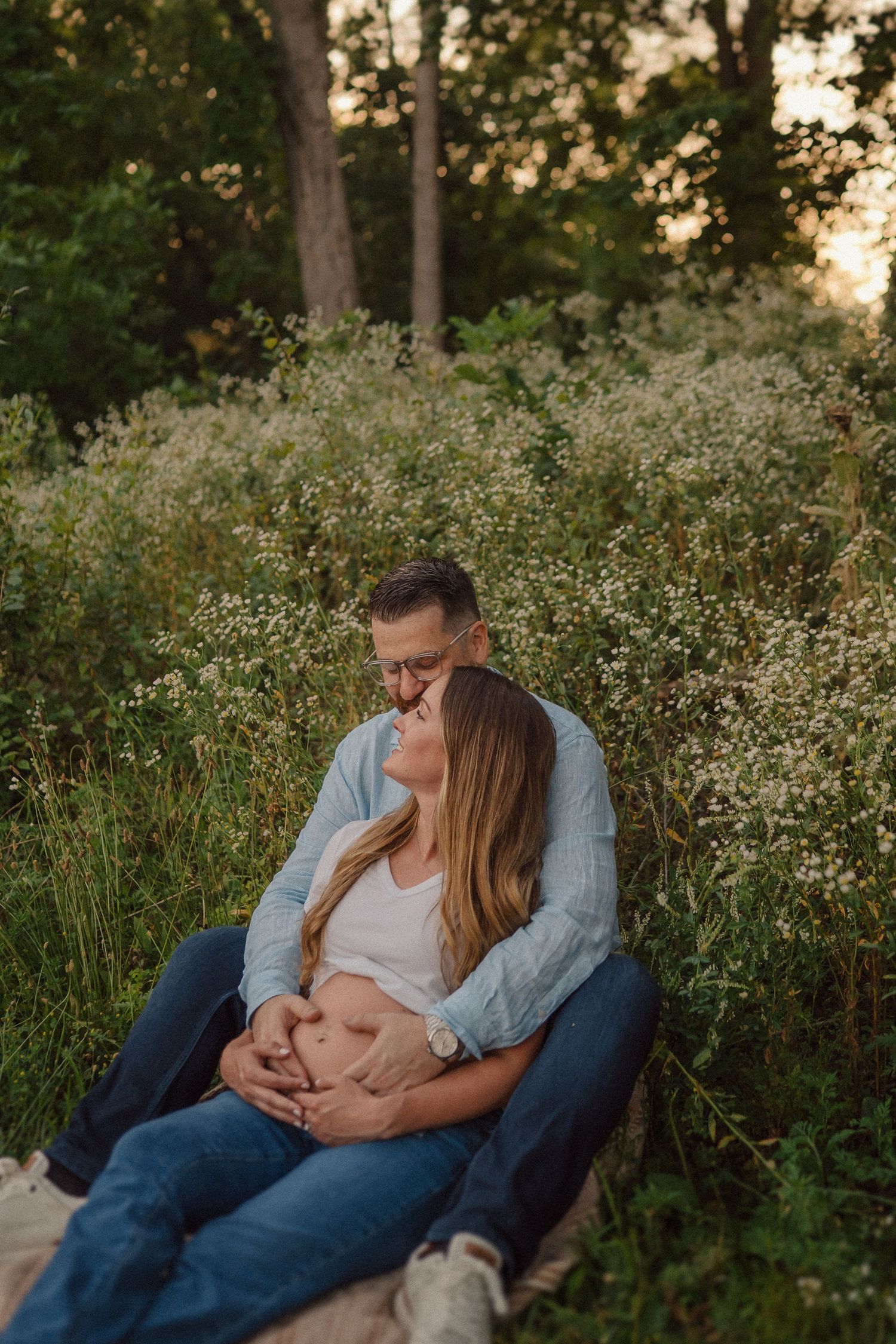 A tender maternity photo shows an intimate outdoor moment with wildflowers and trees during golden hour sunset.