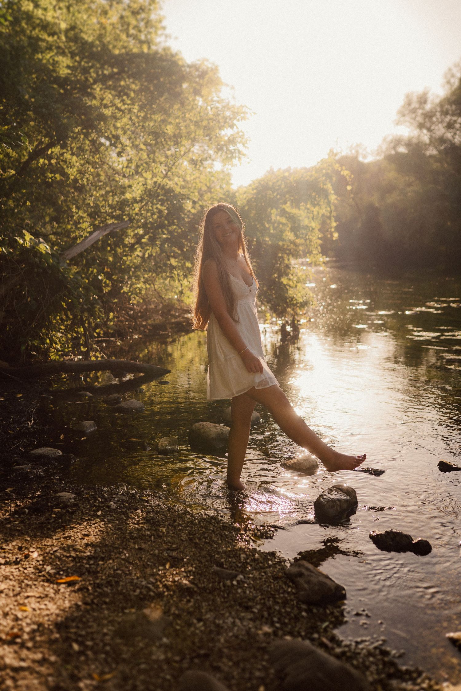 Person in a white dress poses on rocks at the edge of a river during sunset.