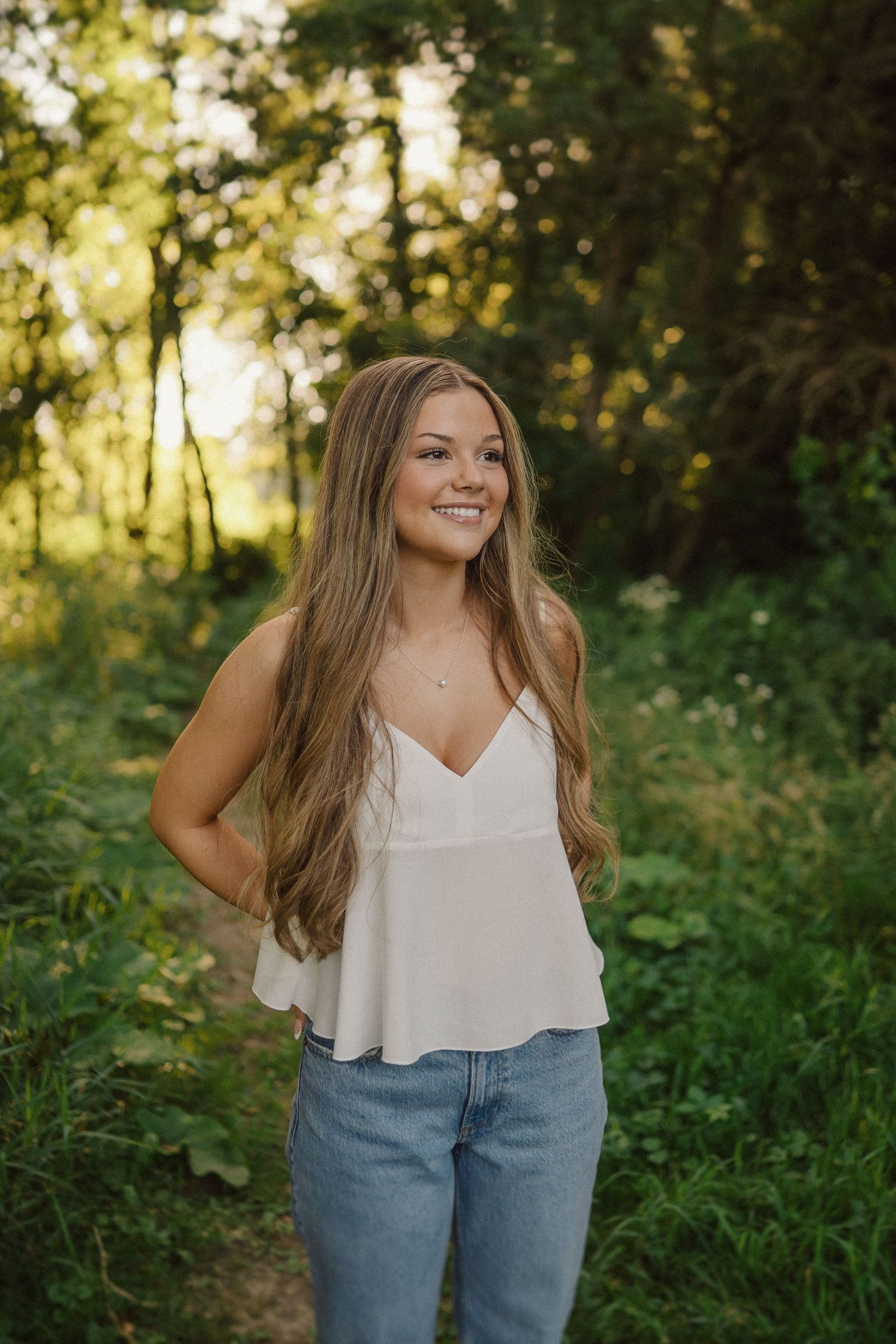 A young woman stands in a forest wearing a white v-neck tank top and light jeans during golden hour lighting.