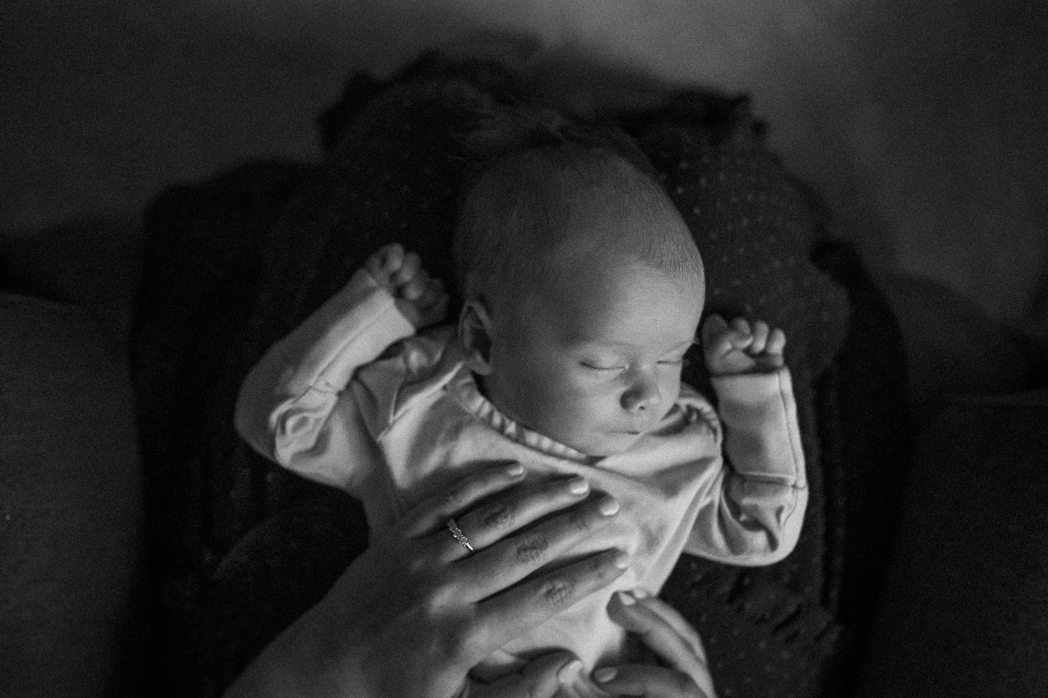 Black and white photo of a sleeping baby nestled in a dark blanket, illuminated by soft lighting.