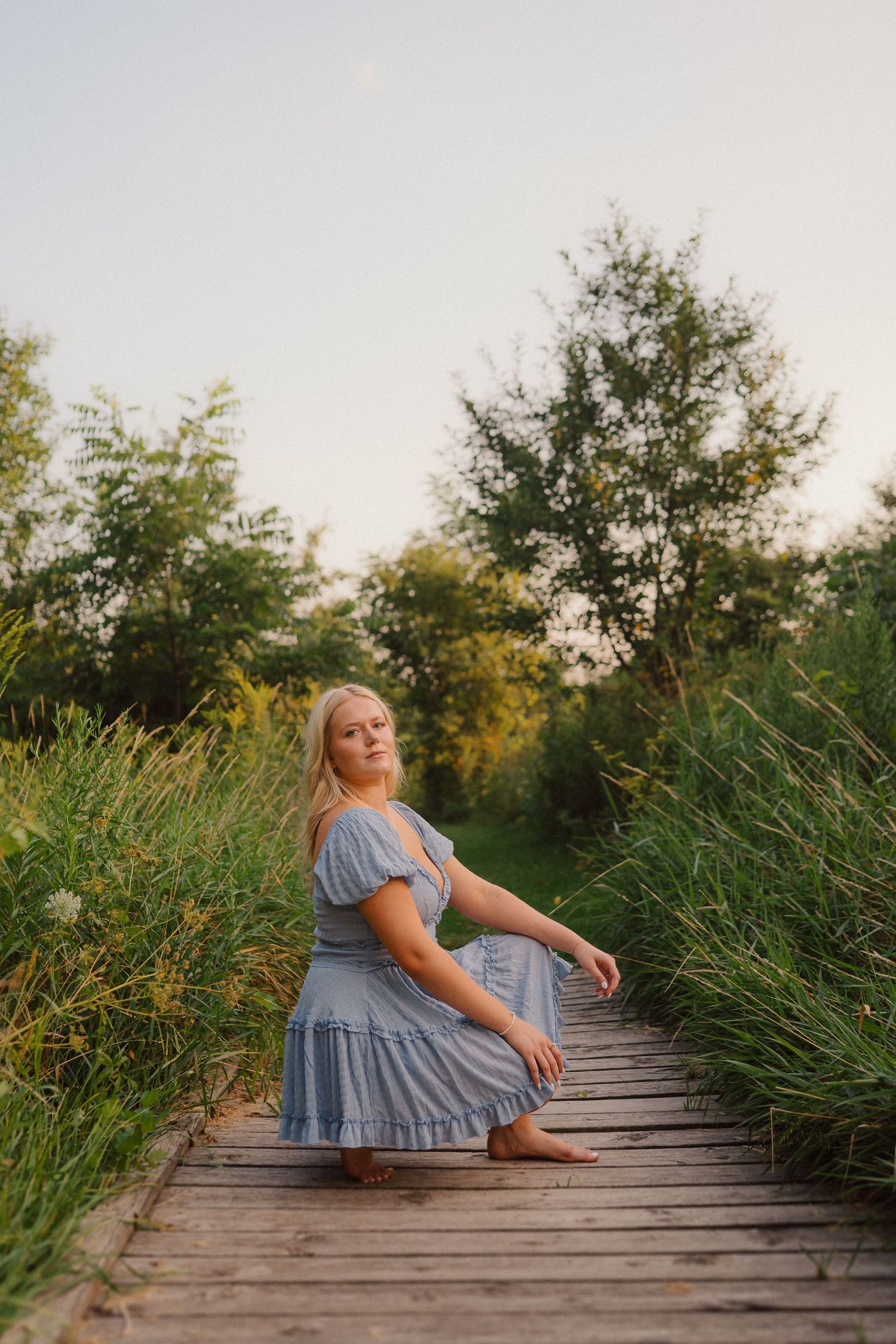 A woman in a flowy blue dress sits elegantly on a wooden boardwalk surrounded by tall reeds at sunset.