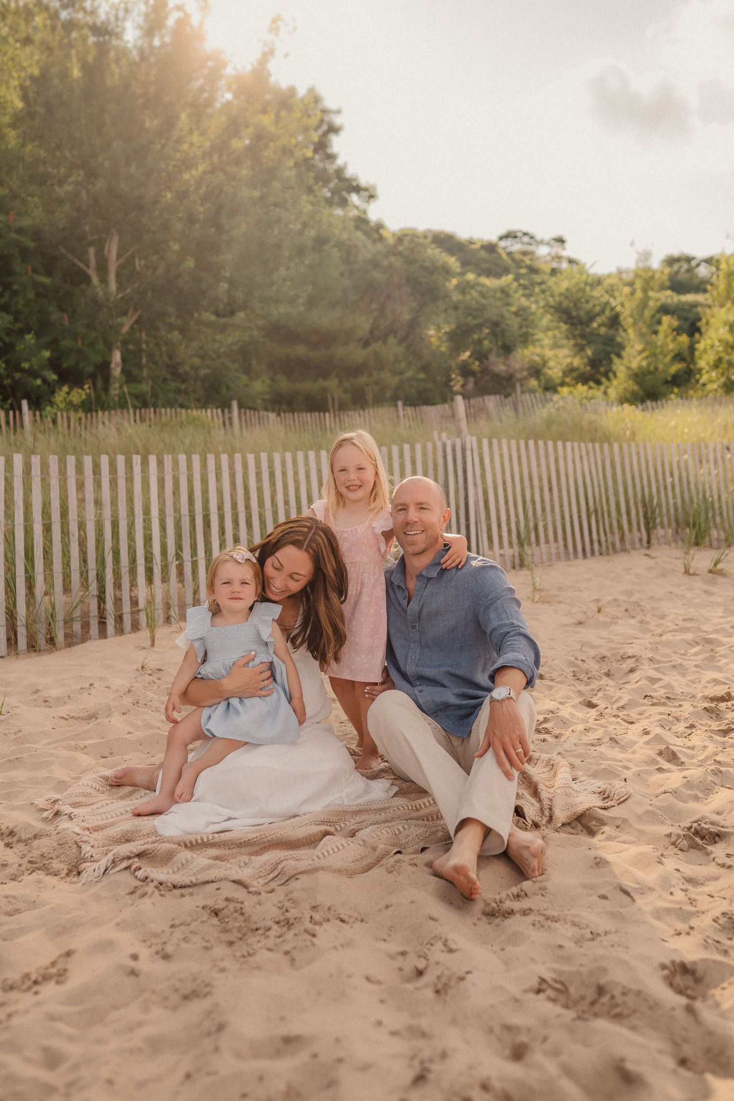 Family members relaxing together on a sandy beach with wooden fence and golden sunlight in background.