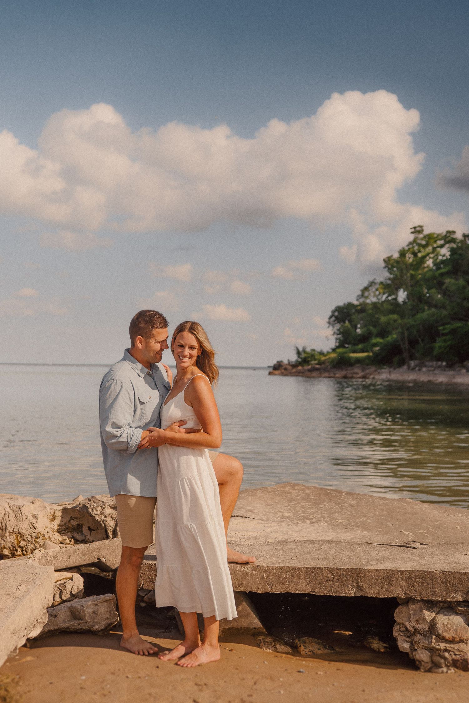 A couple shares an intimate moment on rocky beach overlook during sunset with water views.