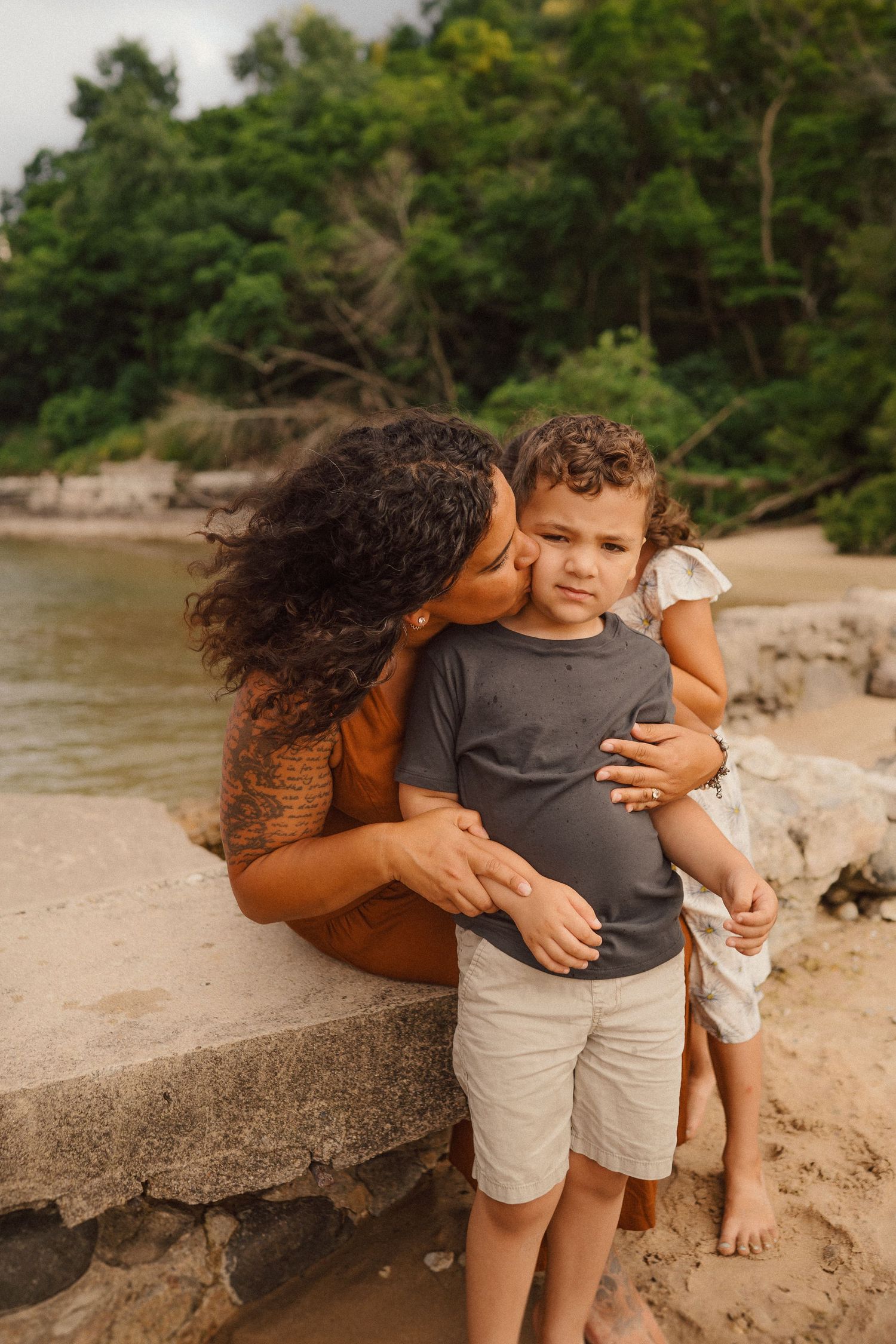 A tender embrace shared between parent and child on a rocky beach with lush trees in the background.