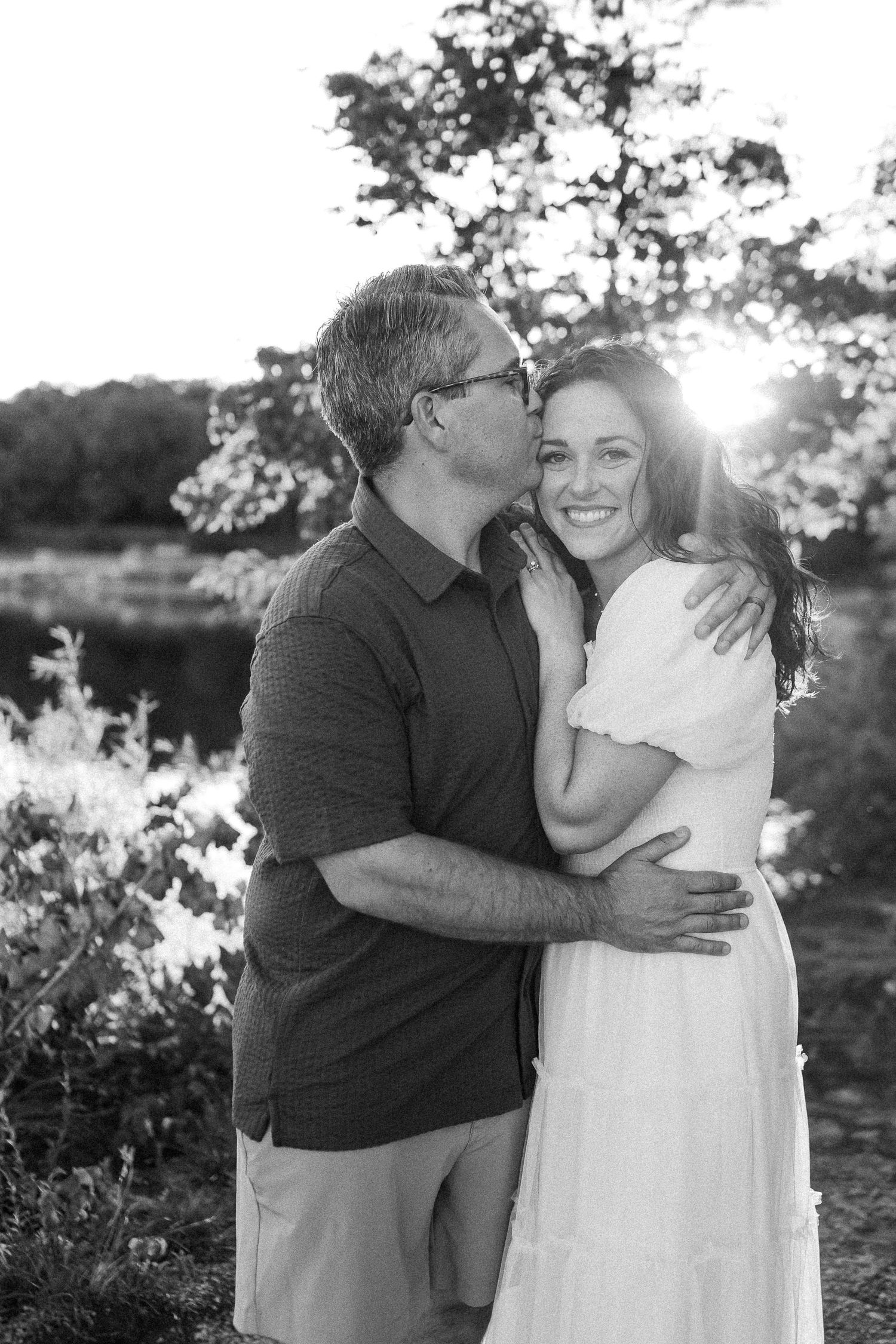 A tender black and white outdoor portrait of a couple embracing in warm sunlight with trees in the background.