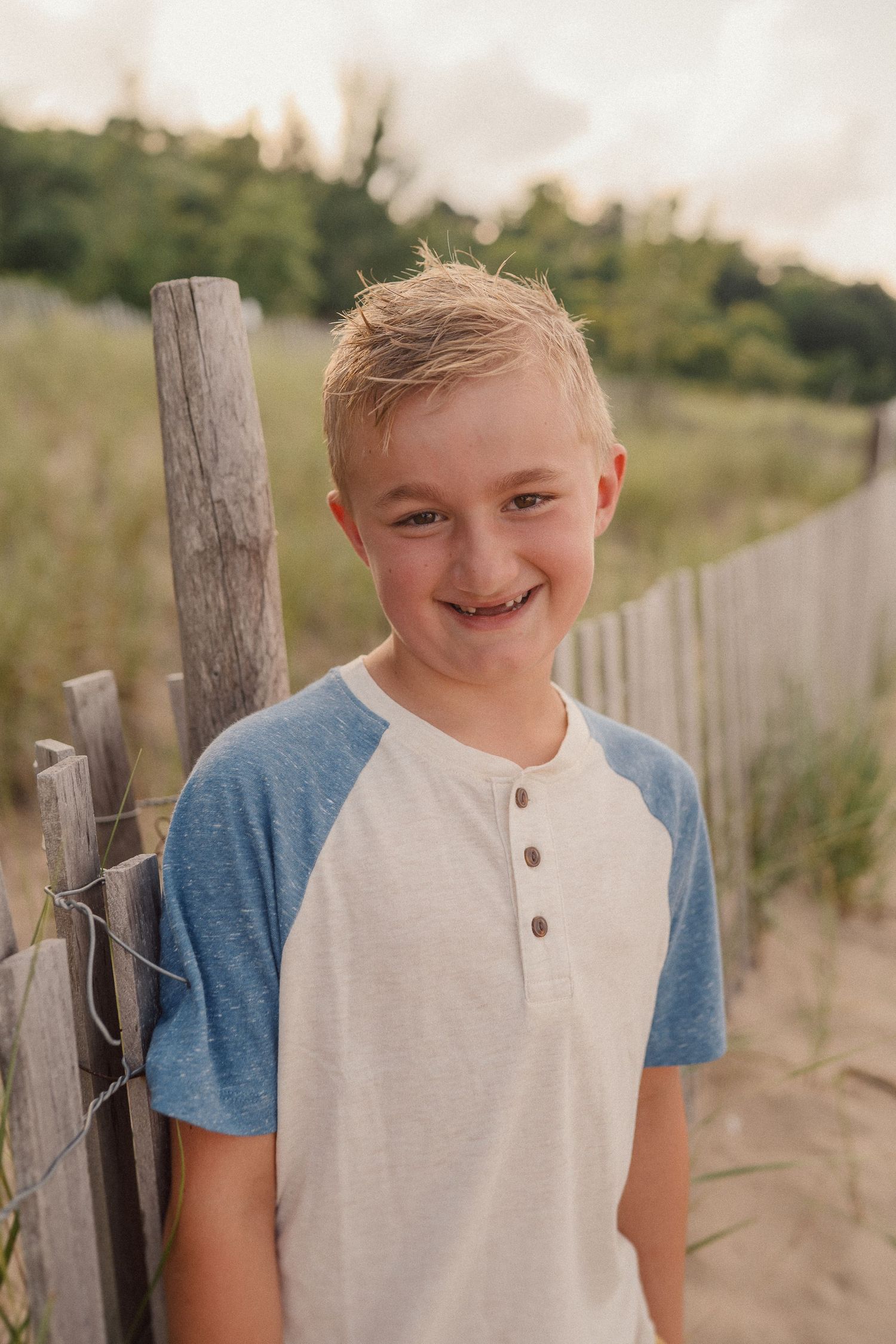 A young person in a blue and white raglan shirt stands smiling next to a wooden fence post on a sandy beach.